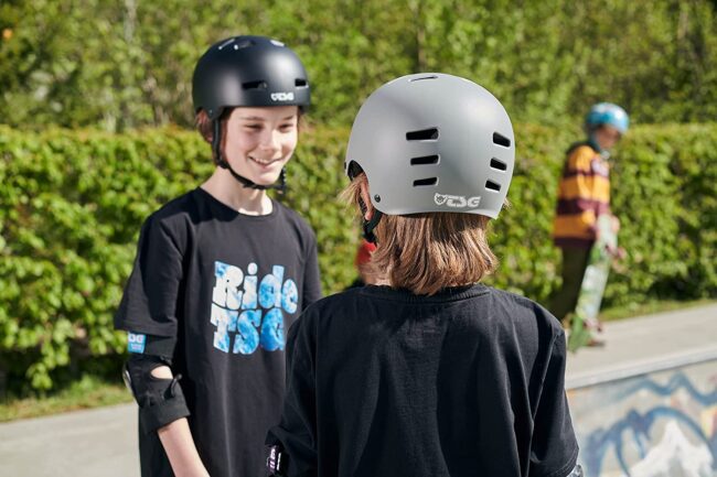 Two young skaters wearing TSG Evolution helmets talking to each other at a skatepark surrounded by greenery.