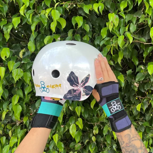 A helmet with a off-white background and colourful flower graphics, held up by hands wearing matching wrist guards against a lush green background.
