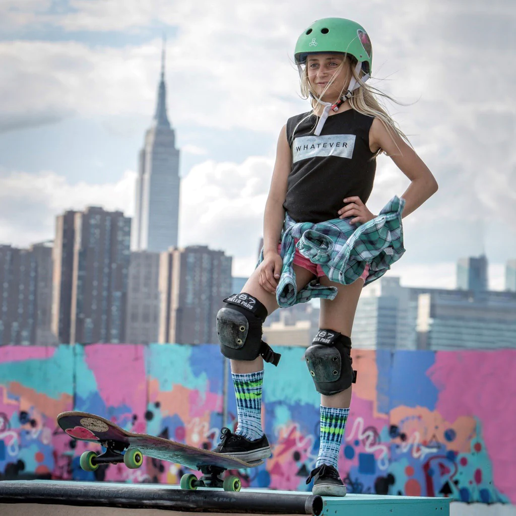 Young female skater about to drop in on a halfpipe, wearing a green Triple 8 helmet, 187 knee pads, and colourful skater socks.