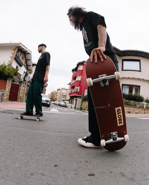 Two skaters in an urban residential area, one preparing to jump onto Globe Goodstock complete skateboard.