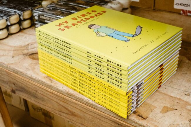 A stack of "My First Skateboard" Volume 1 and "Jonas Meets Jack" books combined, with Spitfire wheels visible behind them on a workbench.