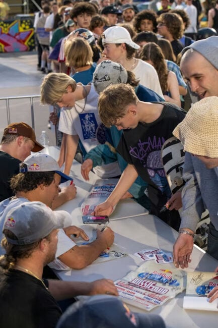 Professional skateboarders, including Jake Wooten, Alex Sorgente and Jamie Foy sitting at a table, signing autographs, and meeting fans. A line of excited people eagerly waits to meet their skateboarding heroes.