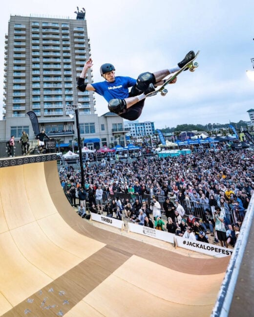 Tony Hawk performs a Stalefish grab mid-air on a large vert ramp at a demo while a crowd of excited spectators watches below.