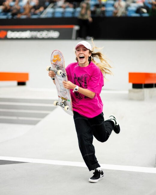 In a lively scene from the SLS event in Sydney, Chloe Covell runs toward the camera with ecstatic energy. Her skateboard raised in celebration, Chloe wears a vibrant pink t-shirt, black baggy skate pants, and Nike SB shoes. The image captures the essence of her excitement and passion for the sport.
