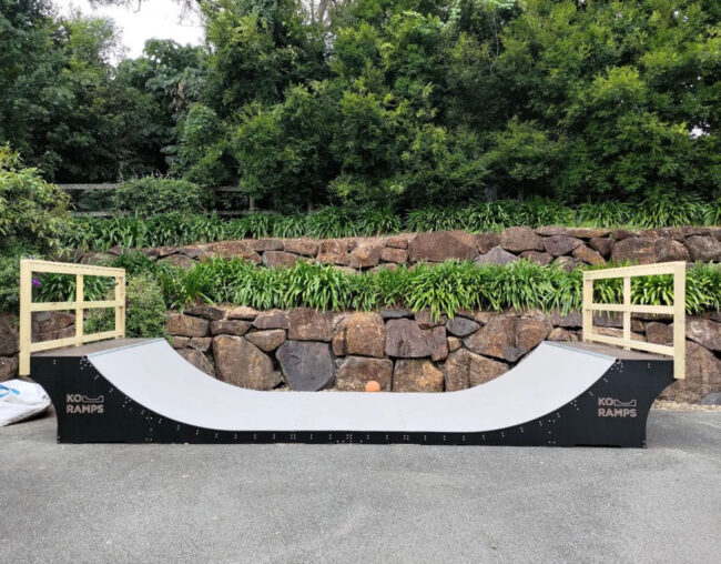 Modern backyard oasis: Ko Ramps' 3-foot high halfpipe with clean lines, dark sides, light grey Skatelite surface, and raw timber handrails. A stylish and updated design blending seamlessly into the picturesque setting.